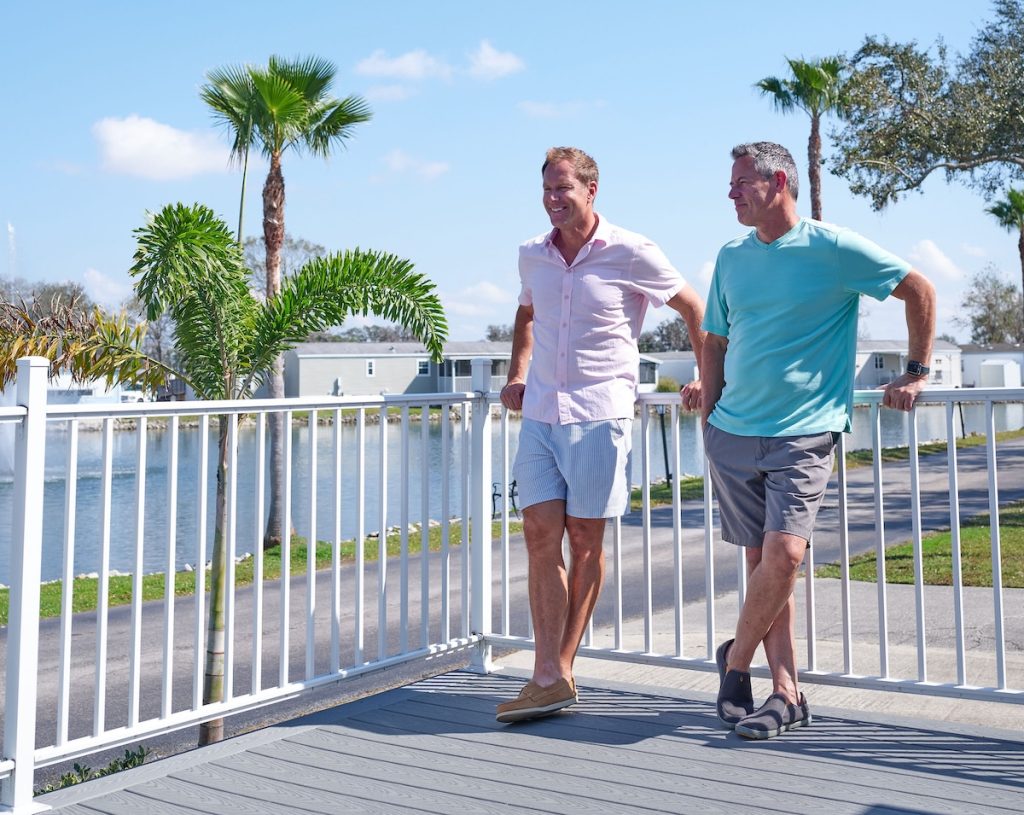 Two men standing on a deck, one in a pink shirt and shorts, the other in a blue shirt and gray shorts, with a view of palm trees and a lake in the background.