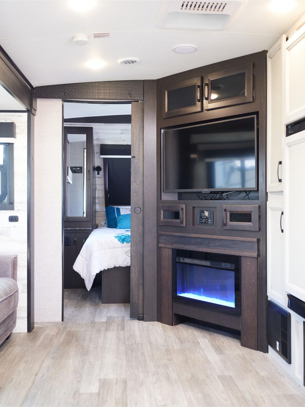 Interior of an RV featuring a wall-mounted TV above an electric fireplace and a view into a bedroom with a bed and white linens.