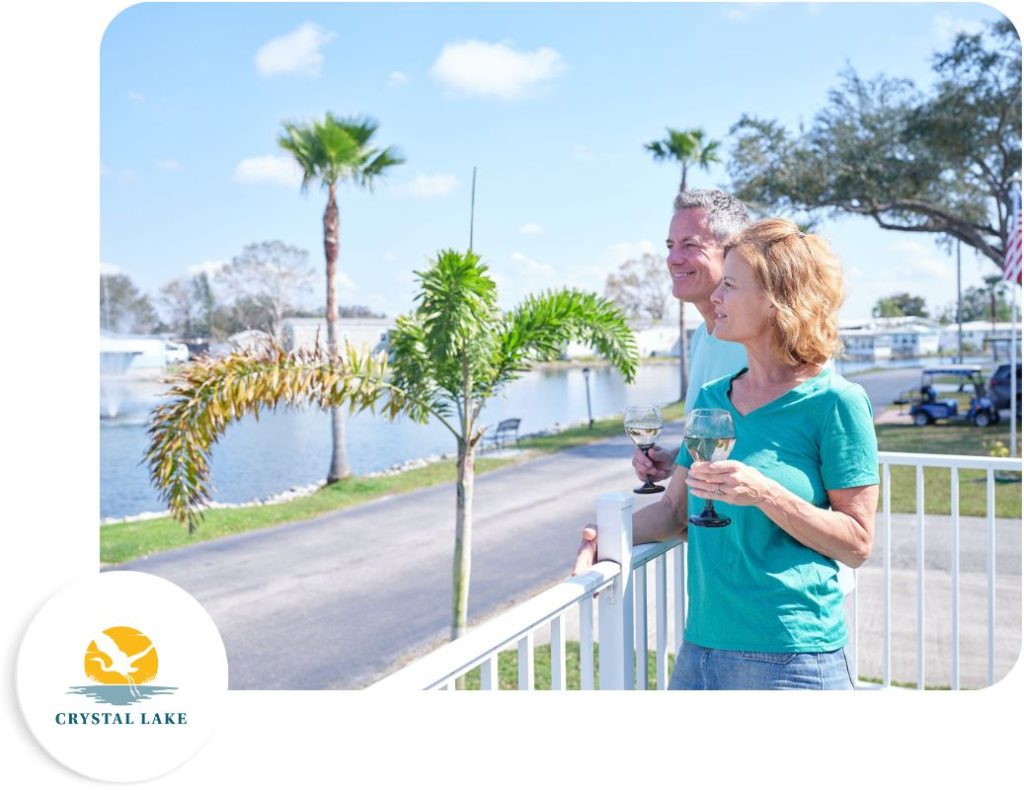 A man and woman stand on a balcony overlooking a lake with palm trees, holding wine glasses. The sky is clear and sunny. There's a "Crystal Lake" logo in the bottom left corner.