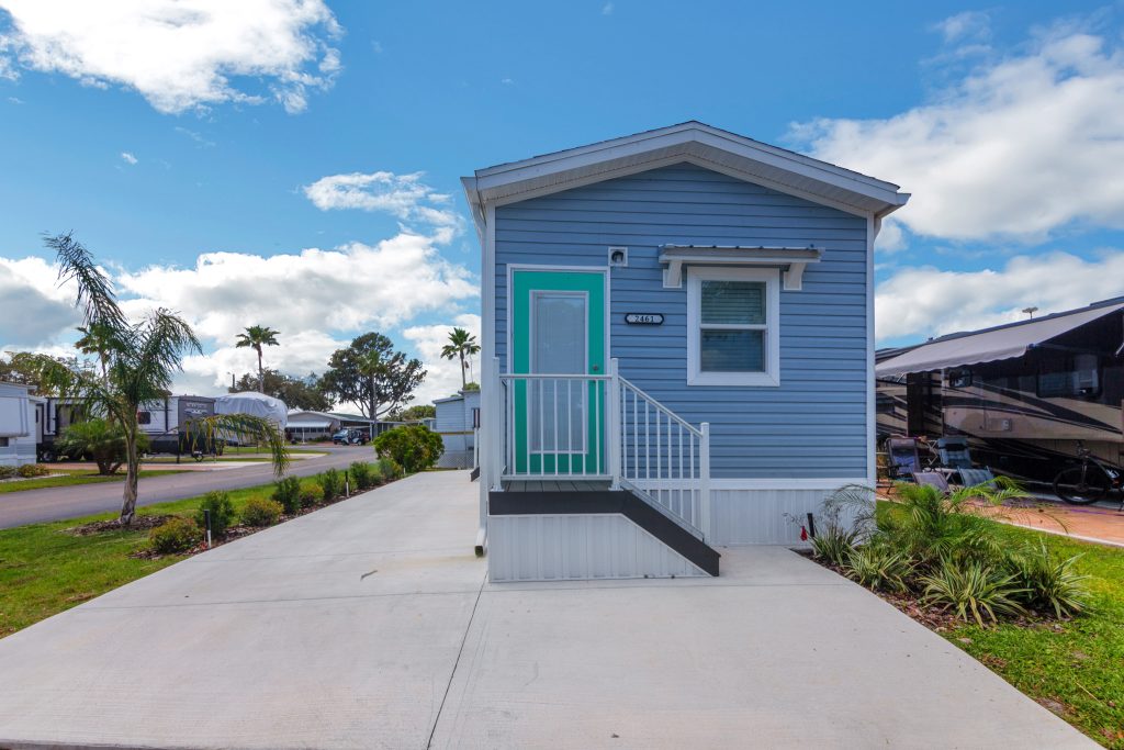 A small, blue mobile home with a green door, concrete driveway, and white railing is situated in a landscaped trailer park under a partly cloudy sky.