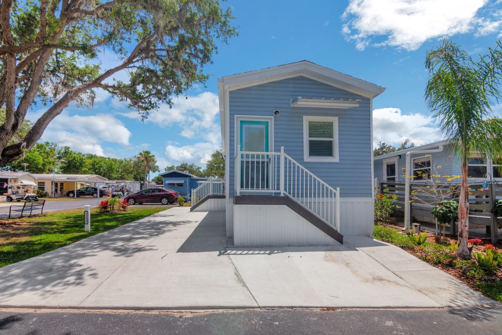 Small blue manufactured home with white trim, front steps, and driveway. Surrounded by trees and neighboring homes under a blue sky with clouds.