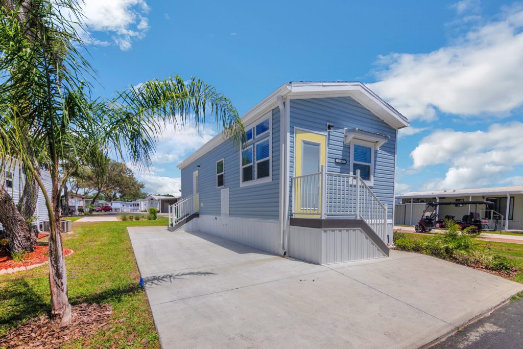 A light blue manufactured home with a yellow door sits on a concrete driveway, surrounded by grass and palm trees under a partly cloudy sky. A golf cart is visible in the background.