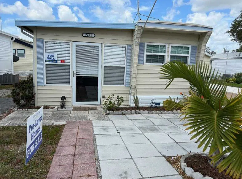 Small beige mobile home with blue trim, front patio, and potted palm. "For Sale" signs displayed on window and yard. White clouds and blue sky in background.