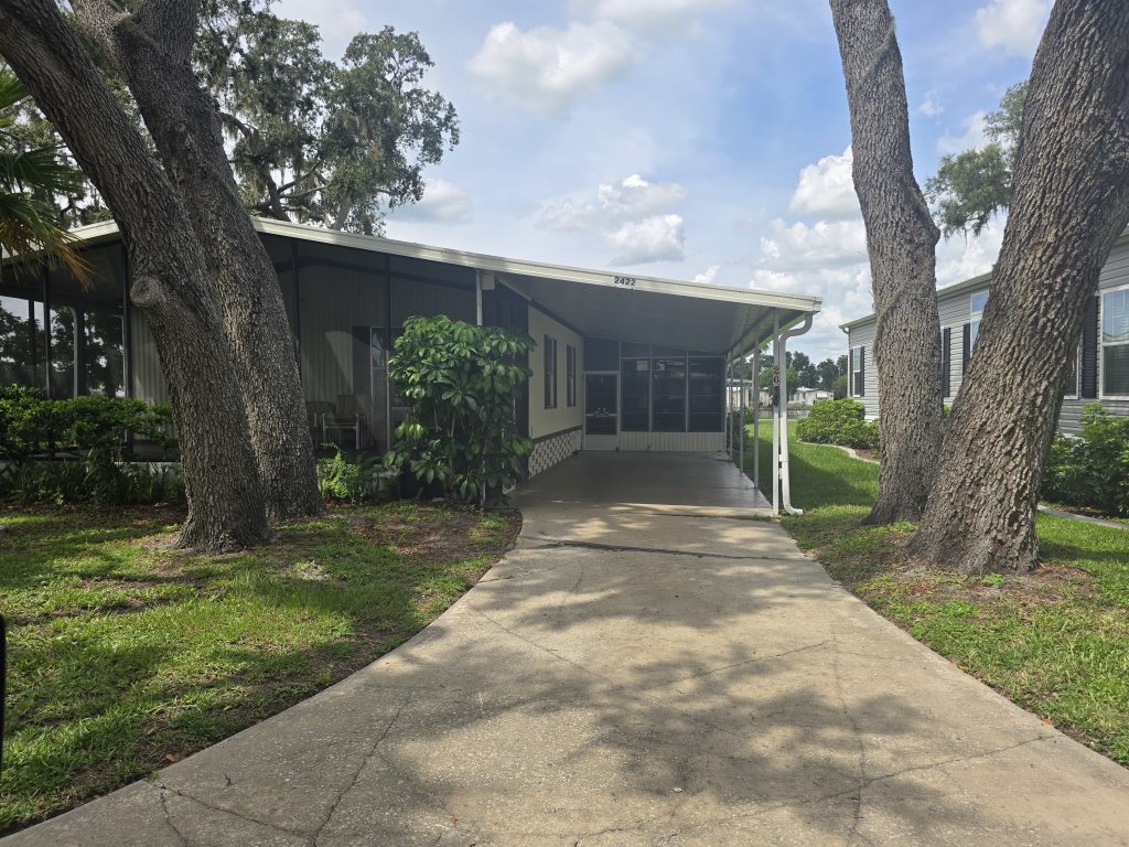 A single-story manufactured home with a covered porch, concrete driveway, and three large trees in the front yard. Neighboring homes and green landscaping are visible.