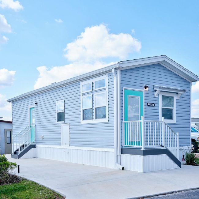 A light blue mobile home with white trim and a turquoise door is set on a concrete pad under a blue sky with clouds.